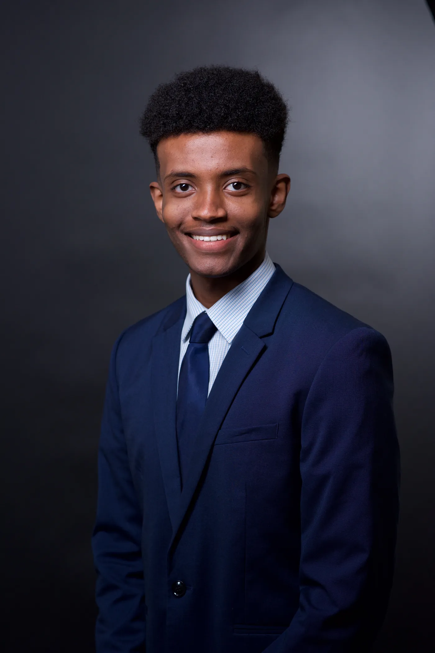 Corporate headshot of a young man in a navy suit on a charcoal backdrop